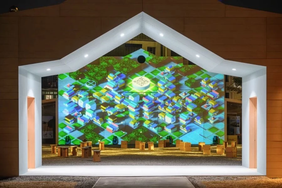 展館位於嘉義市政府對面,整體以方形庭院為核心,其量體造型源自團隊對嘉義木構建築的在地研究。四個導角量體呼應城市街角常見的咖啡館與餐館;四個立面的屋頂線條,則取材自具代表性的木構建築,如博物館與派出所。三側設有通往庭院的入口,室內的粉彩色調亦呼應原始建築的色彩語彙,使其成為一處友善而開放的城市客廳。