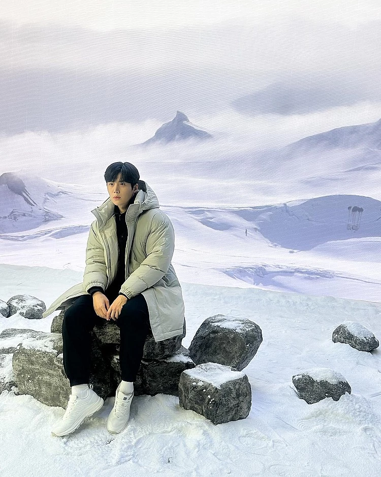 來到雪景場景，金宣虎選擇以淺灰色長版羽絨外套作為造型主角，柔和色調在一片白雪中顯得格外清爽，也成功中和羽絨外套原本容易顯得厚重的視覺感受。內搭刻意全黑配置，讓整體輪廓更俐落，視覺重心自然回到羽絨外套的線條與比例。腳上搭配白色運動鞋，呼應雪地氛圍之餘，也讓造型多了一點輕盈與年輕感。