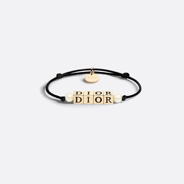 D-Dice 手環讓 Dior 的經典 Logo 從秀場延伸到手腕上。以黑色棉繩搭配金色立體字母骰子，中間鑲嵌白色珍珠，既俏皮又優雅。