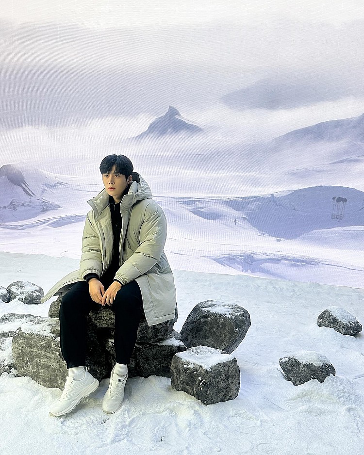 來到雪景場景,金宣虎選擇以淺灰色長版羽絨外套作為造型主角,柔和色調在一片白雪中顯得格外清爽,也成功中和羽絨外套原本容易顯得厚重的視覺感受。內搭刻意全黑配置,讓整體輪廓更俐落,視覺重心自然回到羽絨外套的線條與比例。腳上搭配白色運動鞋,呼應雪地氛圍之餘,也讓造型多了一點輕盈與年輕感。