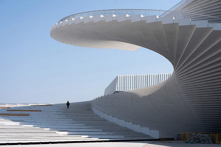 坐落於上海市浦東新區世博文化公園內，一座重量級新建築——上海大歌劇院——即將登場，並由挪威建築事務所 Snøhetta 操刀設計。臨河而建的位置、大量純淨的白色表面，以及彷彿自地面緩緩升起的平緩建築輪廓，構成其整體風貌。最具標誌性的元素，是一座違反重力感受的螺旋樓梯。這座形似菊石的樓梯承擔更宏觀的美學功能，使整個建築彷彿以其為核心，如扇面般向外展開。