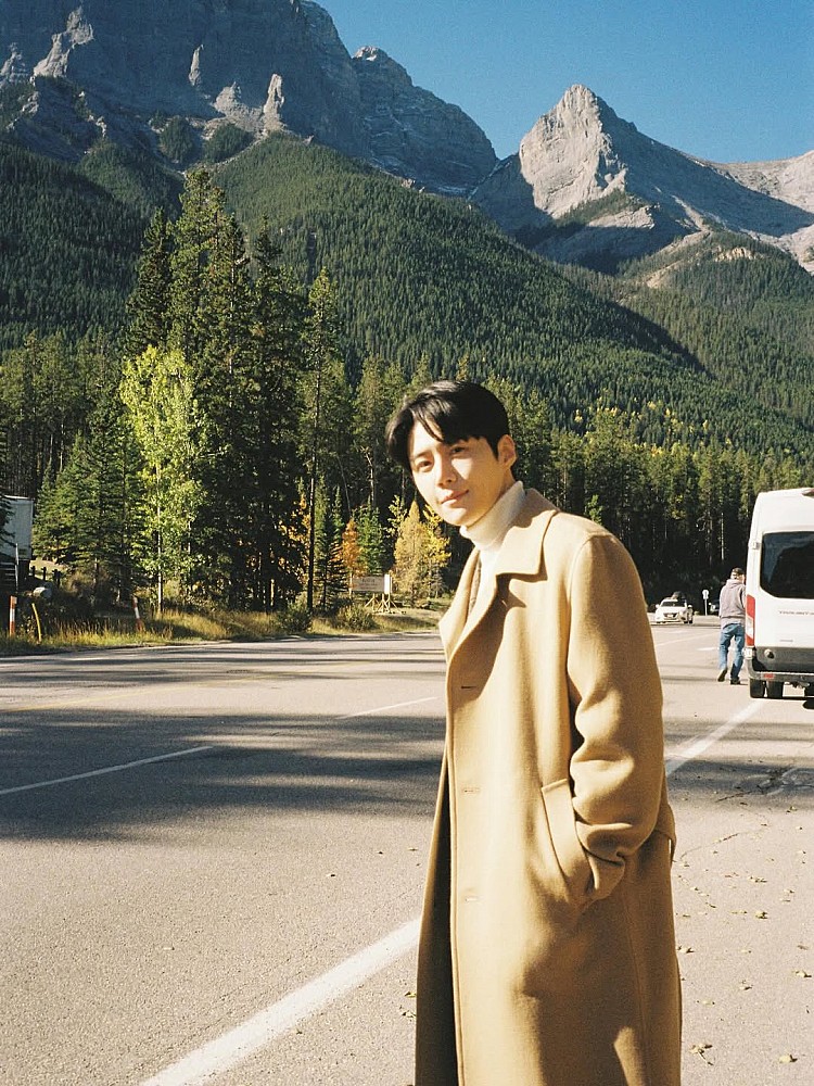 這一套造型裡，金宣虎選擇了淺駝色長版毛呢大衣作為主角，顏色溫潤、不搶戲，卻在自然光下顯得格外耐看。內搭一件白色高領針織，利用乾淨的色彩對比，讓整體視覺更加明亮，也自然拉出臉部線條。