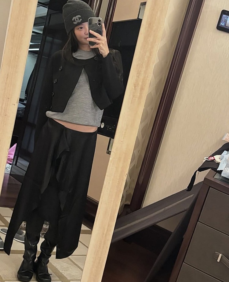 Jennie以灰色針織上衣搭配不規則剪裁長裙，展現鬆弛又高級的極簡穿搭。柔軟針織貼合身形，外搭短版黑色外套拉長身形比例，巧妙平衡俐落與慵懶。