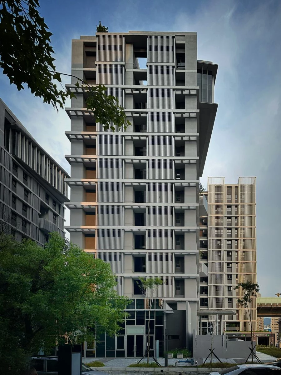 位於台中大里溪舊河道的重劃區內，育賢二期社宅以延展地景紋理為設計主軸，使量體自然融入河道脈絡。公共廊道串接周邊街廓與一至三期基地，於地面層形塑具公共共享性與社會互動性的開放空間。立面以一致的模矩語彙建立秩序感，讓建築與周遭街區呈現緊密且協調的城市關係。