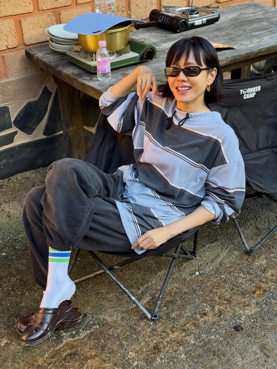 這套戶外休閒造型，是孫藝珍私服中編輯私心最喜歡的一套。微高領設計的寬鬆條紋上衣，搭配深灰色束口棉褲，本身就帶有一點90年代的輕復古氣息；再加上黑色長方形墨鏡與踩腳皮鞋，讓整體造型立刻有了重心，從單純的休閒感，往更「有造型意識」的方向推進。腳邊若隱若現的藍綠條紋白襪，巧妙替下半身增添層次與亮點，讓造型多了一點玩心，卻不顯刻意。這樣的搭配不強調曲線，也不追求修身，而是透過比例與細節取勝，呈現出一種自在、不費力卻很有風格的時髦感。