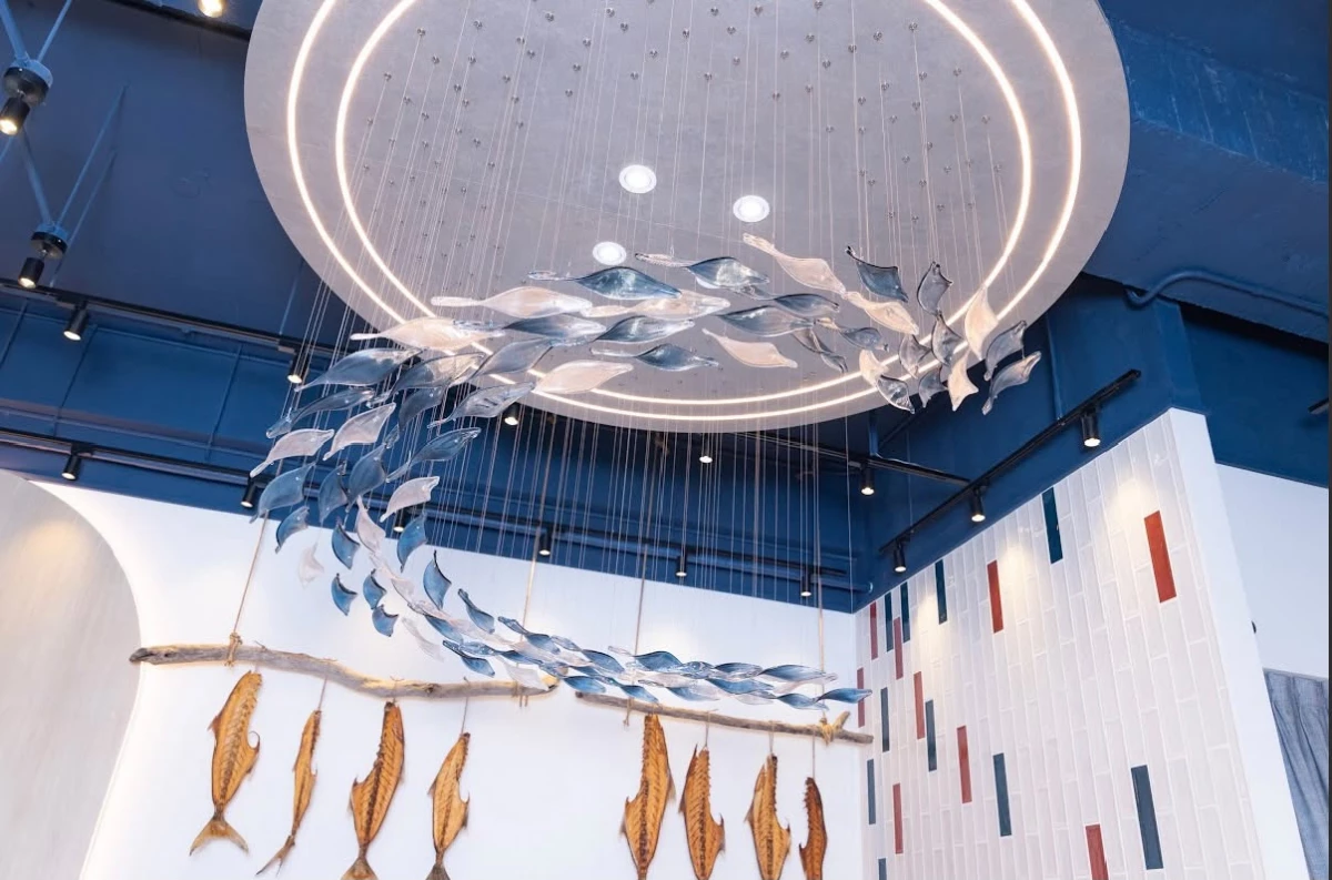 除了料理，空間也是 FISH BAR 250° 品牌體驗的重要一環，走進店內映入眼簾的是懸掛於中央的大型魚群藝術裝置，透明與深藍色魚體沿著圓形燈帶盤旋，就像看著魚群隨著海流游動，天花板以深海藍為主色，搭配溫潤木質、石紋檯面與白色牆面，在冷暖色調之間建立平衡。