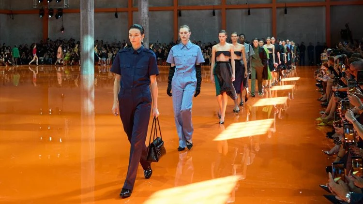 Prada 2026 春夏大秀於Fondazione Prada的Deposito空間揭幕，整個場域化作一片耀眼的橘色，地板覆上高亮塗層，映照如鏡般的光澤，營造出虛實交錯的奇幻氛圍。