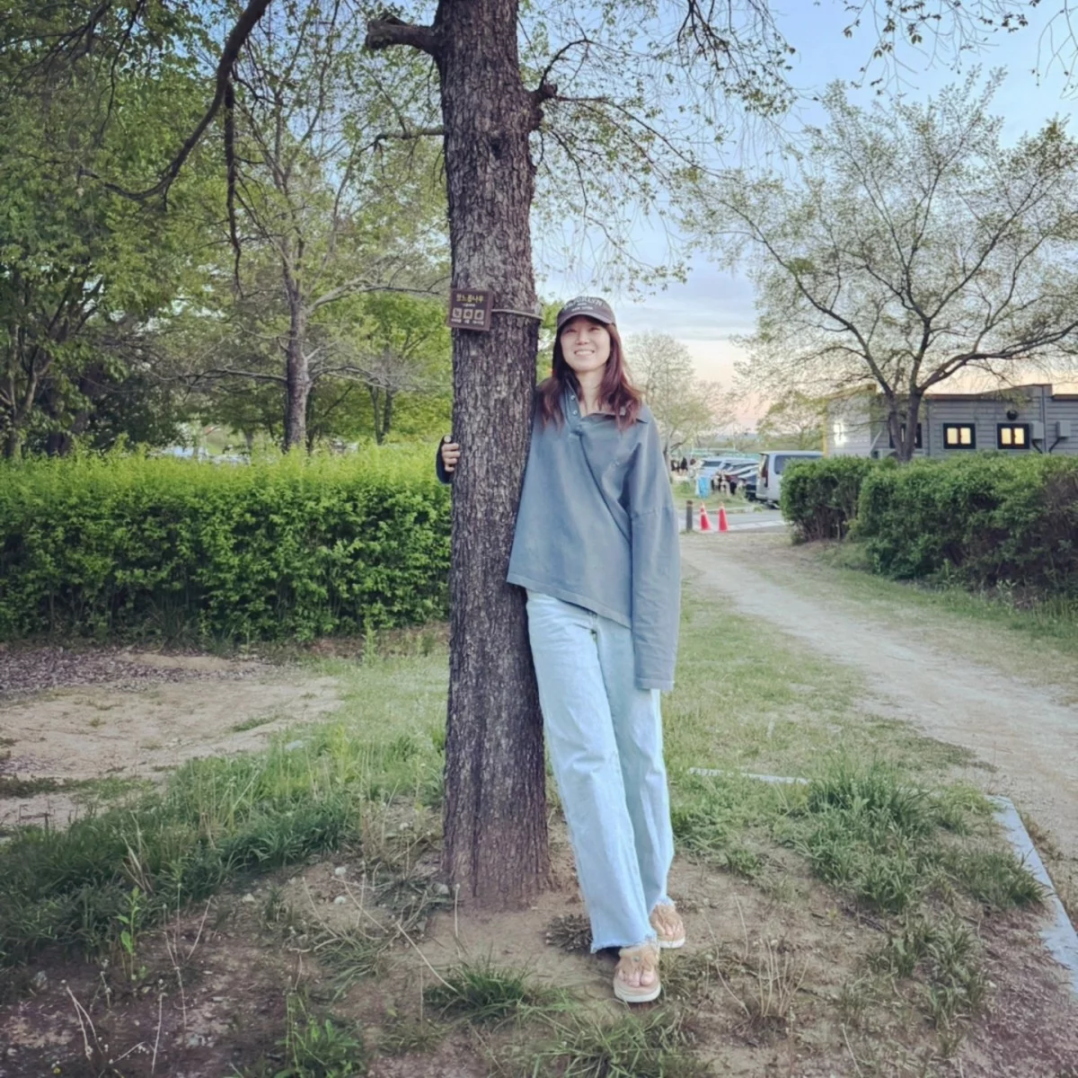 孔曉振以灰色 oversized 亨利領上衣搭配淺色寬版牛仔褲，戴上棕色棒球帽在露營地靠著大樹拍照