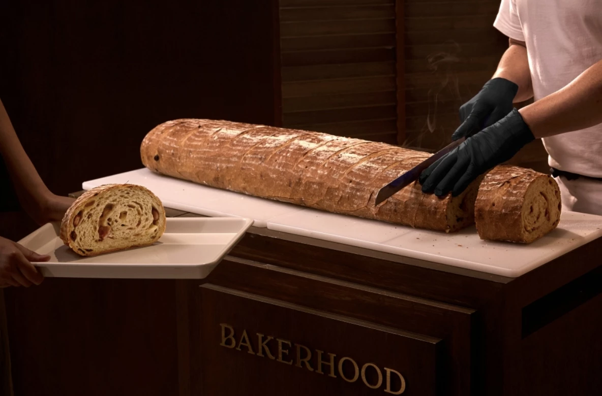 選址於充滿生活感的靜謐巷弄，BAKERHOOD 以麵包為策展主題，揉入麥香、綠意與職人精神，打造一處讓現代人遠離塵囂的新場域。其中最吸睛的莫過於品牌 Logo 中出現的招牌長棍麵包：長達 100 公分的「普羅旺斯暢遊」。這款超長歐法麵包每日現場切片販售，內包裹德國香腸、培根、油漬番茄與雙色橄欖，不僅視覺吸睛，也展現品牌分享與交流的精神。
