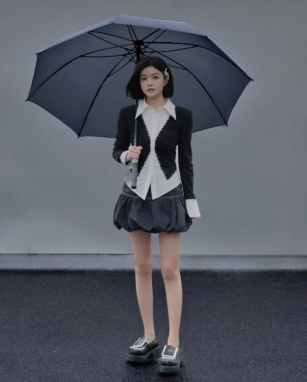 這套雨中街拍走的是完全不同的氣場路線。陳妍希身穿黑白撞色鉚釘西裝外套,內搭白襯衫,下身配黑色泡泡裙,短鮑伯頭加上黑色大傘,整體造型帶點復古歐美時裝感。腳上的黑色厚底樂福鞋同樣是「餅乾鞋」家族的一員,銀色釦環細節呼應外套上的鉚釘元素,厚實的鞋底讓泡泡裙的蓬鬆感不會顯得頭重腳輕,反而撐出更挺拔的比例。這套證明厚底鞋不只能穿出減齡少女感,走氣場、走個性路線同樣適用。