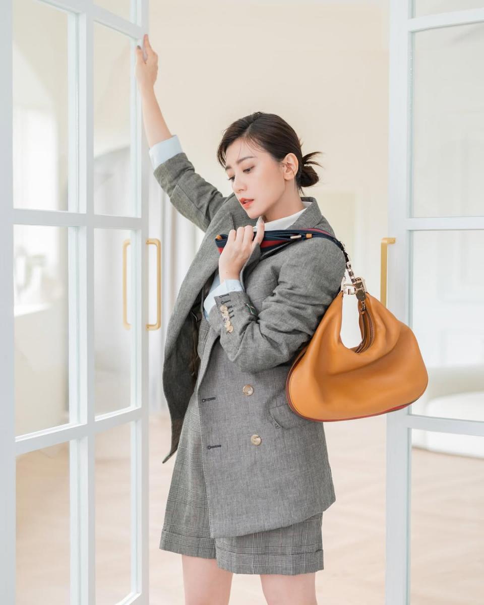 灰色西裝向來給人理性、正式的印象，但賈靜雯這套造型成功把它穿得輕鬆不老氣。她選擇線條俐落卻不貼身的西裝套裝，透過微寬鬆的比例，讓整體輪廓保有呼吸感，也避免過度成熟。造型關鍵落在包款的選擇，她手拎 Gucci Attache 琥珀色肩背包，搭配品牌經典撞色背帶，為偏中性的灰色調注入一抹溫度與時尚識別度。半月型的大包不只實用，也柔化了西裝的剛性線條，讓整體看起來更慵懶時髦。這樣的搭配不刻意追求年輕，而是透過比例與配件，讓西裝自然走進日常，是非常聰明的穿搭示範。