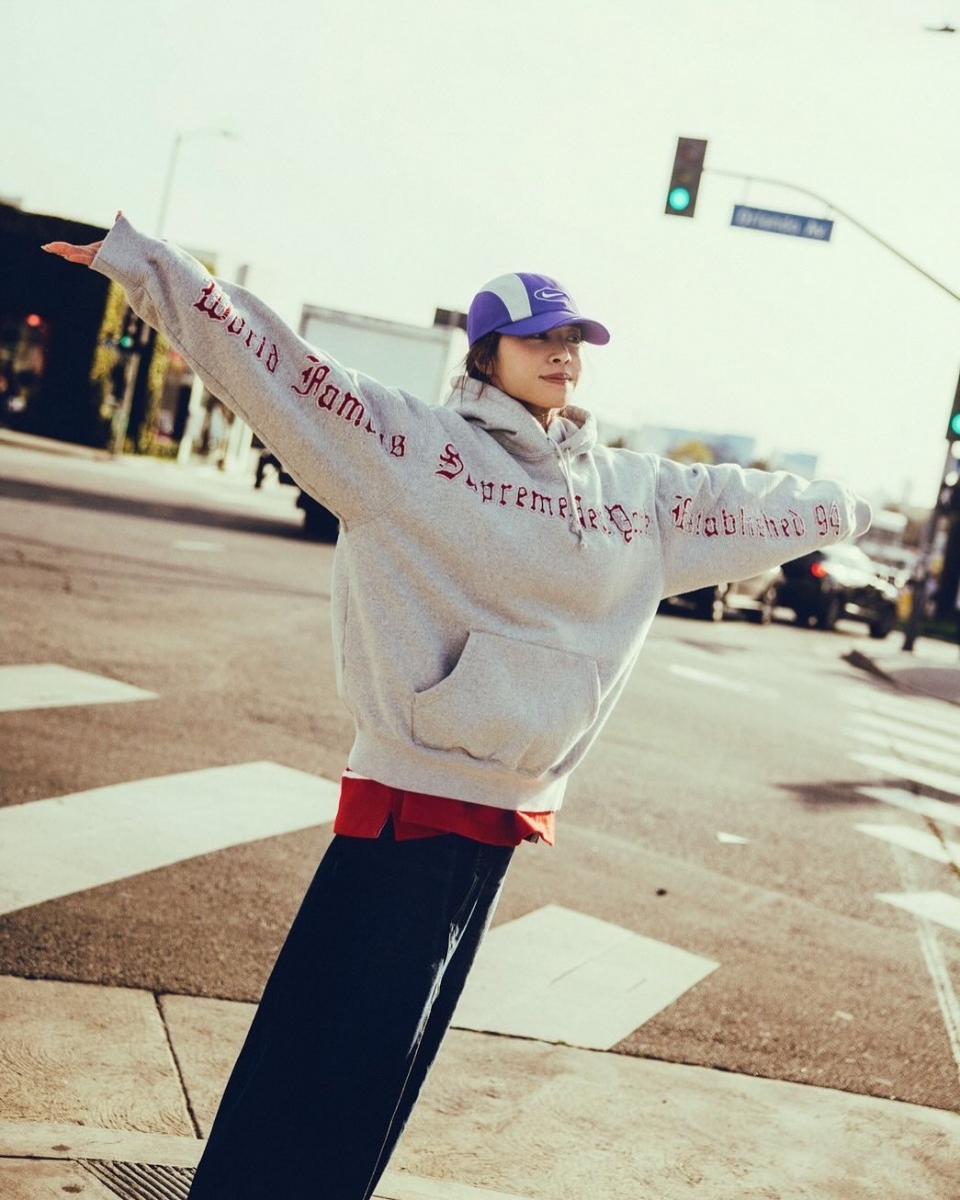 這套造型把街頭風的「玩色精神」拿捏得剛剛好。灰色字母帽T作為主體，份量感寬鬆卻不顯拖沓，替整體奠定隨性基調；下擺刻意露出的紅色內搭，像是不經意的一筆亮色，除了呼應連帽上衣印刷的英文字母色調外，也讓造型瞬間有了節奏與層次。紫色棒球帽則成為視覺焦點，為整身注入年輕、帶點叛逆的街頭氣息。這樣 Oversized 的中性穿搭不追求展露身材曲線，而是利用顏色與比例營造隨性自由的風範，穿出一種不被年齡定義的從容態度，自然顯得年輕又有型。