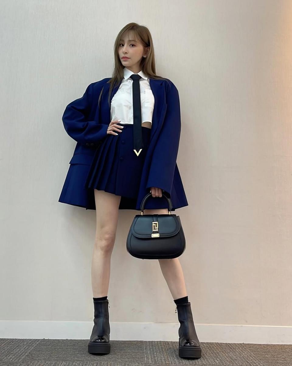 這套造型把「正式感」轉化為顯高利器。寶藍色套裝以一致色調建立完整縱向線條，視覺集中、不被切割，本身就有拉長比例的效果；內搭選擇短版白襯衫，刻意露出腰線位置，讓上短下長的結構一目了然。領帶元素與手上的 Versace Greca Goddess 迷宮女神包，進一步加強垂直延伸感，讓視線自然由上而下移動。下身搭配厚底短靴，不僅補足高度，也為整體造型增加份量感。王心凌這套穿法證明，只要比例拿捏得當，小隻女也能穿出「甜中帶酷」的俐落風範。